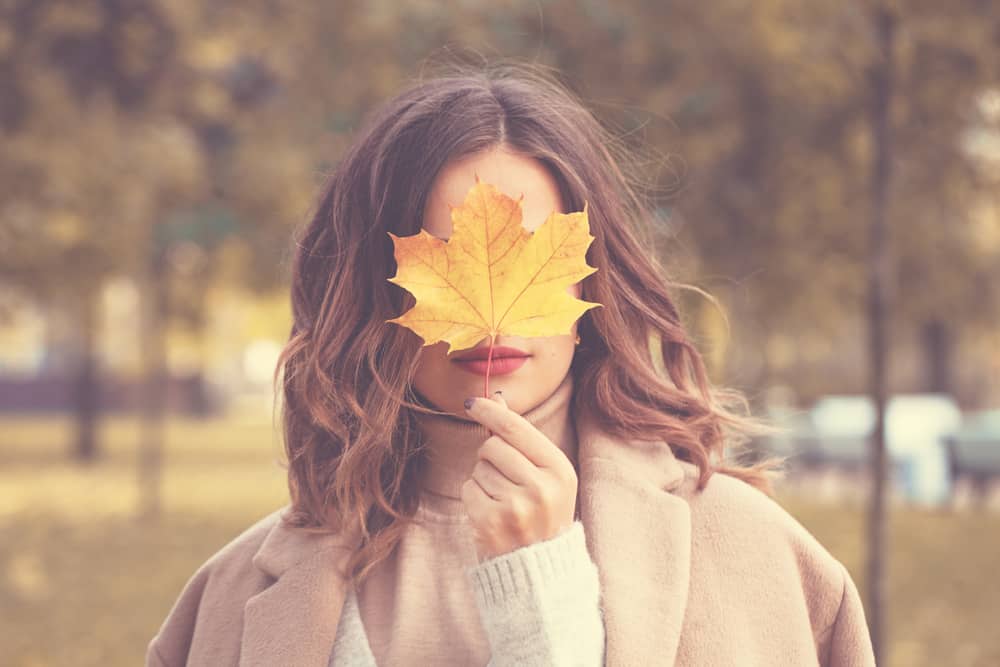 fall image of a woman in brown hair standing in a field wearing a tan jacket and holding a yellow leaf in front of her mouth