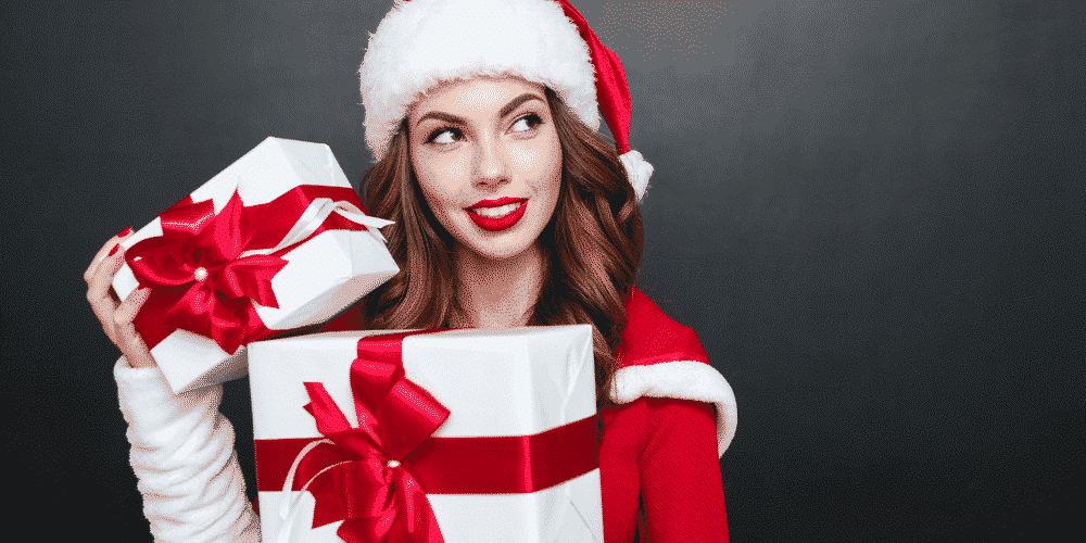 woman with red lips dressed in santa outfit holding white presents with red ribbons while looking to the side